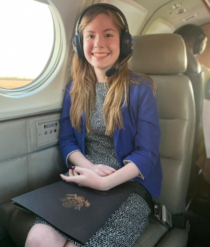 A woman sitting in an airplane, wearing headphones. She has long hair and is smiling.