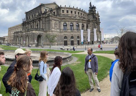 People gathered around a speaker in front of a byzantine building