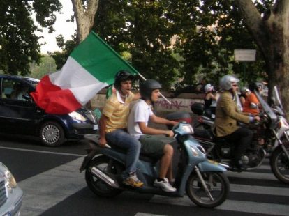 Two guys riding a moped while holding Italian flag.